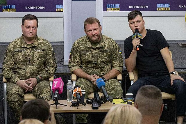 Three men in military uniforms and one man in civilian clothes sitting at a table with microphones.
