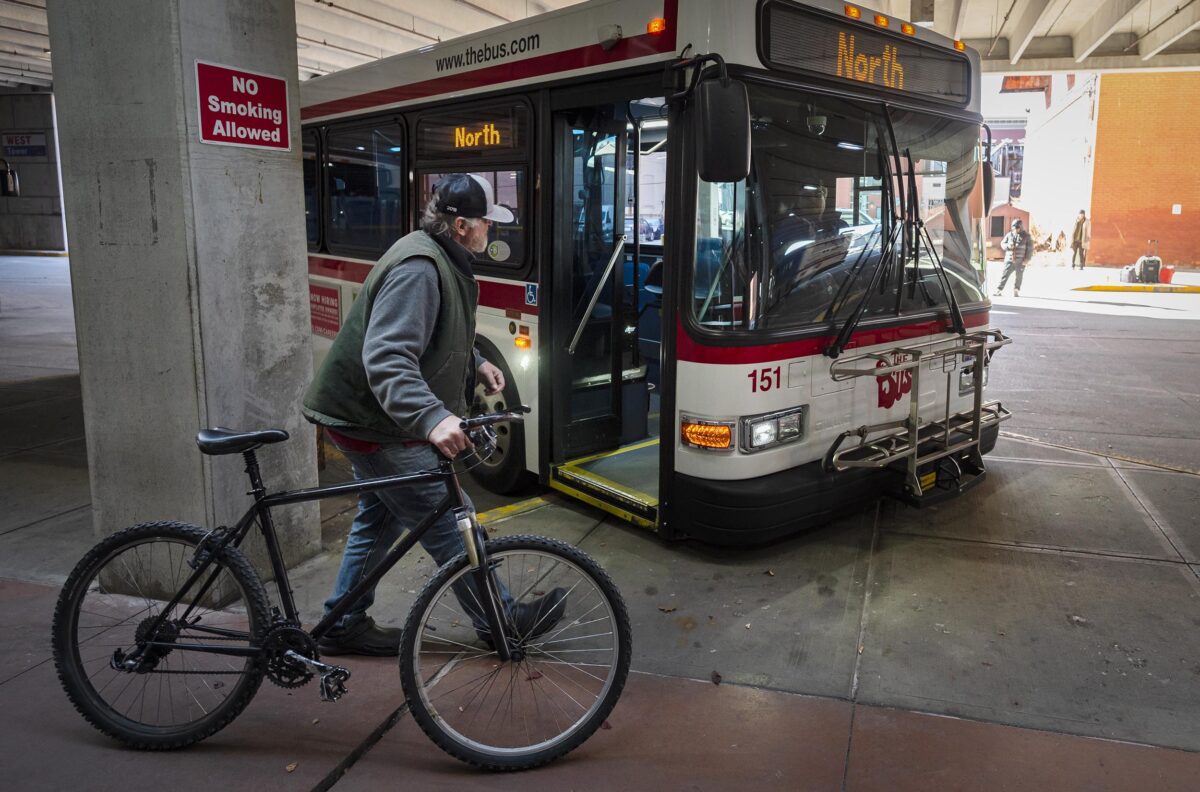 Man with a bicycle approaches a public bus marked "North" at a bus terminal. A "No Smoking Allowed" sign is visible.