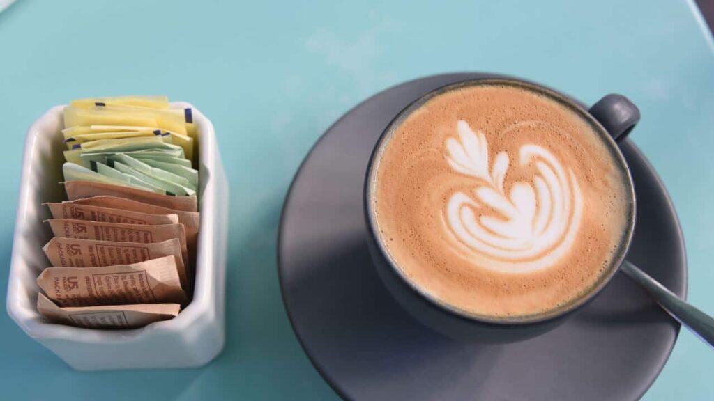 A cup of coffee on a plate, sitting on a teal coloured table. A container with packets of sweeteners is next to it.
