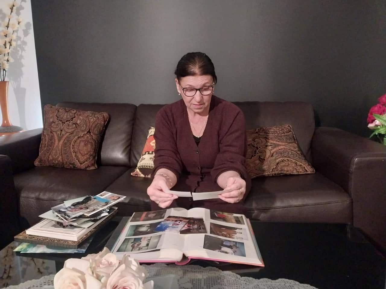 An older woman with dark hair and dark rimmed glasses, wearing a brown cardigan, sitting on a brown sofa looking at a photo album.
