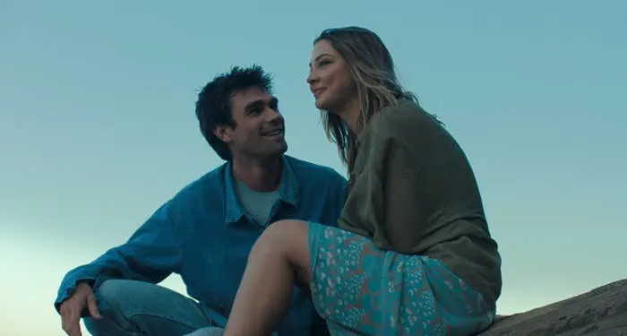 A couple smiles at each other, sitting on rocks by the sea. The woman wears a floral skirt and shawl; the man wears a casual shirt and jeans