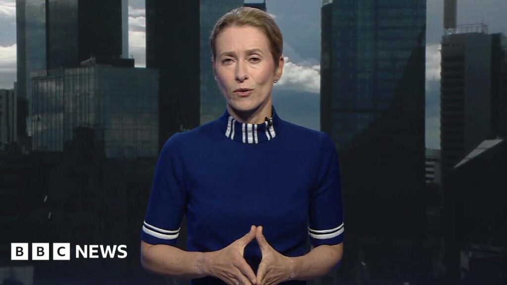 A blonde woman in a high-necked blue top is speaking to a camera, with a microphone clipped to her collar. Her hair is tied back and she is gesturing with her hands pressed together.