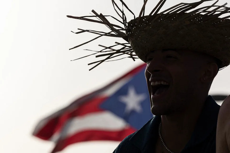 A man wears a straw hat with the Puerto Rican flag in the background before the start of the first show of Puerto Rican singer Bad Bunny’s 30-date concert residency at the Coliseo de Puerto Rico in San Juan, on July 11, 2025. (Credit: Ricardo ARDUENGO / AFP)