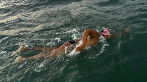 Premal Pattani Premal Pattani wearing black Speedo swimming trunks a red neoprene hat and goggles. He is swimming front crawl in the open sea at dusk, and the water is a deep dark green colour. 