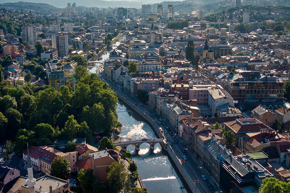 A cityscape showcasing a river winding through buildings, lush greenery, and distant mountains under the sunlight. A mix of modern and historic structures.