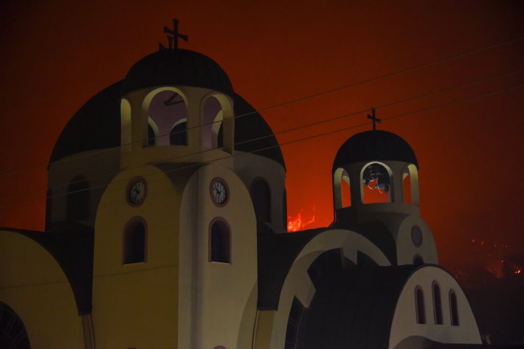 Blazes approach a Greek Orthodox church during a wildfire in the village of Tsoukalaiika near Patras city, western Greece, Tuesday, Aug. 12, 2025.