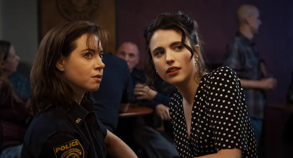 Two women sit closely in a dimly lit room; one in a police uniform, the other in a polka dot dress, both with serious expressions