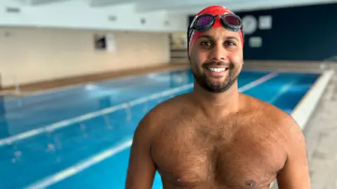 BBC Premal Pattani wearing a red swimming cap and black swimming goggles, smiling at the camera. He is shirtless and standing beside an empty indoor swimming pool, which is out of focus in the background. 