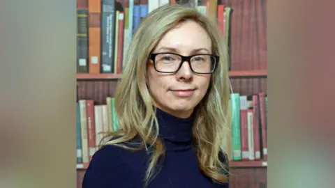 AFBI A headshot of Dr Yvonne McElarney with blurred bars at either side. She has long blonde hair and is wearing a pair of black rimmed glasses. She is wearing a navy turtleneck jumper.
