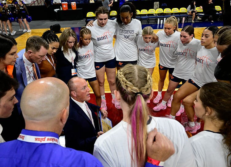 High school girl basketball players stand in a circle around a male coach who's crouching and speaking to them.