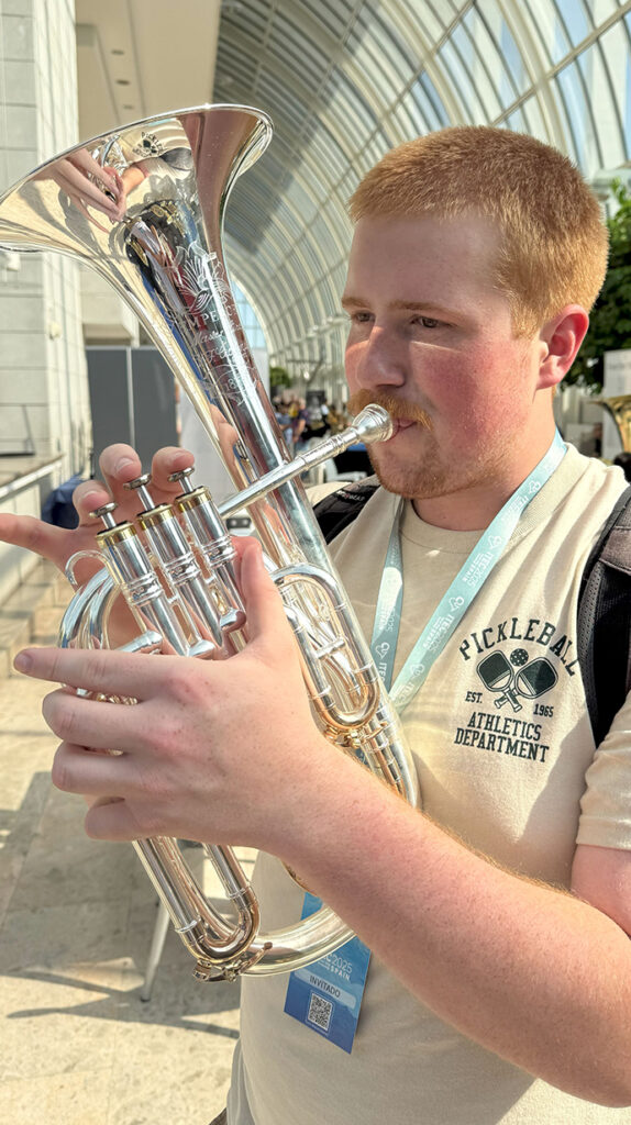 Music Student Hopes Competition in Spain Pays Dividends A young man plays a brass instrument in an atrium with a glass ceiling.