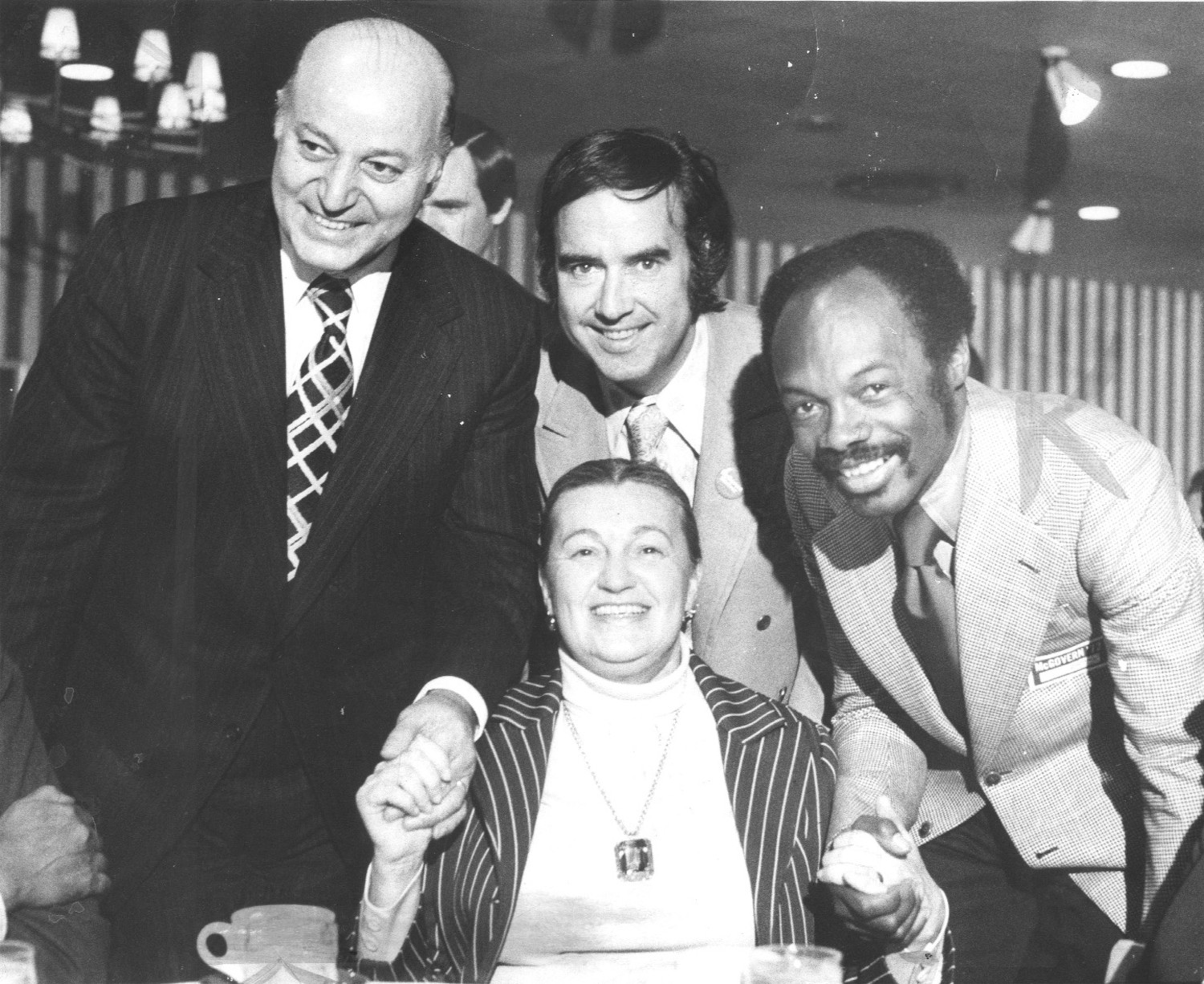 Two men and one woman stand smiling around a seated woman, all dressed formally, holding hands in a warmly lit indoor setting.