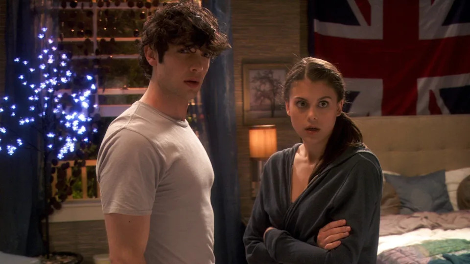 Two people stand in a bedroom, looking surprised. A British flag is visible on the wall behind them
