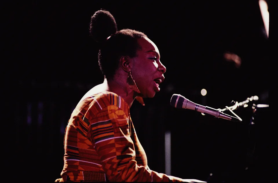 A woman sings passionately into a microphone while playing a piano on stage, wearing a patterned top and large hoop earrings
