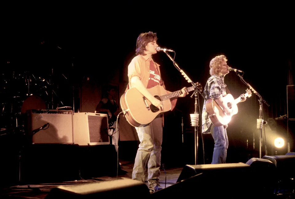 Two musicians perform on stage with guitars, singing into microphones. Stage lights illuminate them as they engage the audience