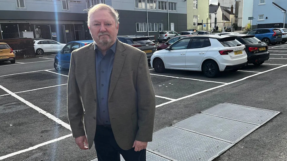 A man looks at the camera. He has a blue shirt and a grey jacket on with black pants. He has wispy grey hair and a grey goatee beard. The bunker can be seen bottom right of the image. As well there are cars that can be seen in the car park.
