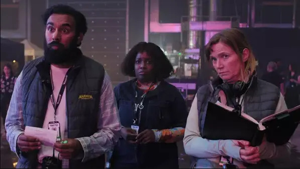 Three people at an event, one holding a notebook. They're wearing casual jackets with badges. The setting appears to be backstage or a media area