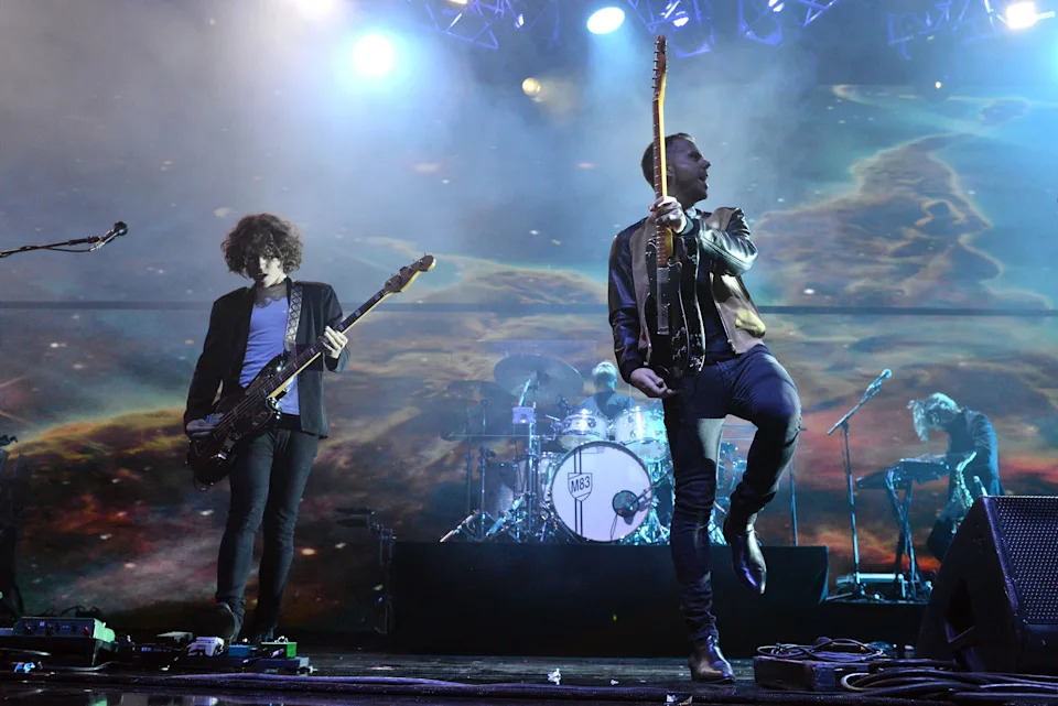 Band performing energetically on stage with guitars and drums, dynamic lighting in the background