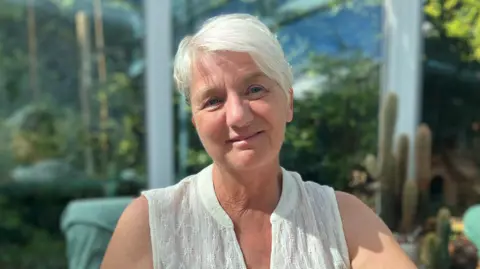 BBC Sandra Nicklin - she has short, white cropped hair and is wearing a white top - a garden can be seen in the background. 
