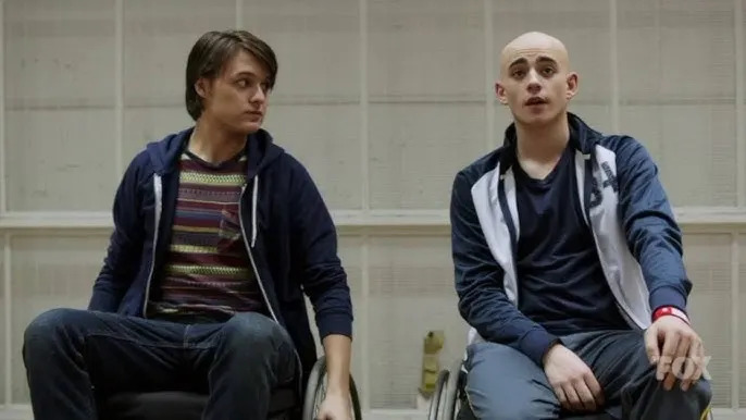 Two people in wheelchairs, one with hair wearing casual shirts and jeans, and one bald wearing a jacket and track pants, talking indoors