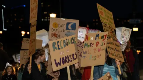 Rob Browne People at a protest at night hold up placards with messages on reading 'recognise us as workers' and 'how many more balls will i have to juggle'.