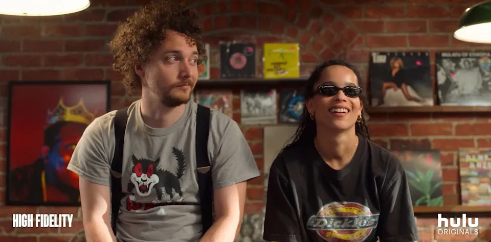Two people sit in a vinyl record store. One wears a graphic tee and suspenders, the other wears sunglasses and a printed tee. Background shows album covers