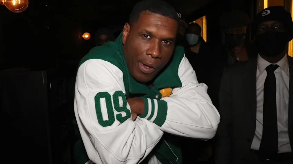 Jay Electronica in a green and white varsity jacket poses with arms crossed. Others in the background wear suits and masks.