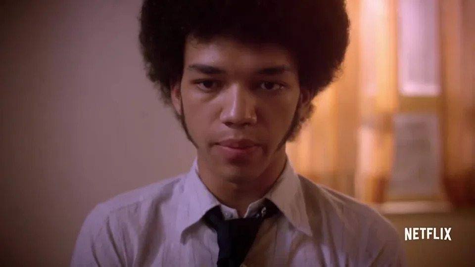 A person with an Afro hairstyle in a formal button-up shirt and tie, looking directly at the camera. Netflix logo in the corner