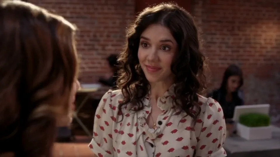 Two people having a conversation in a casual indoor setting. One person has curly hair and is wearing a patterned blouse