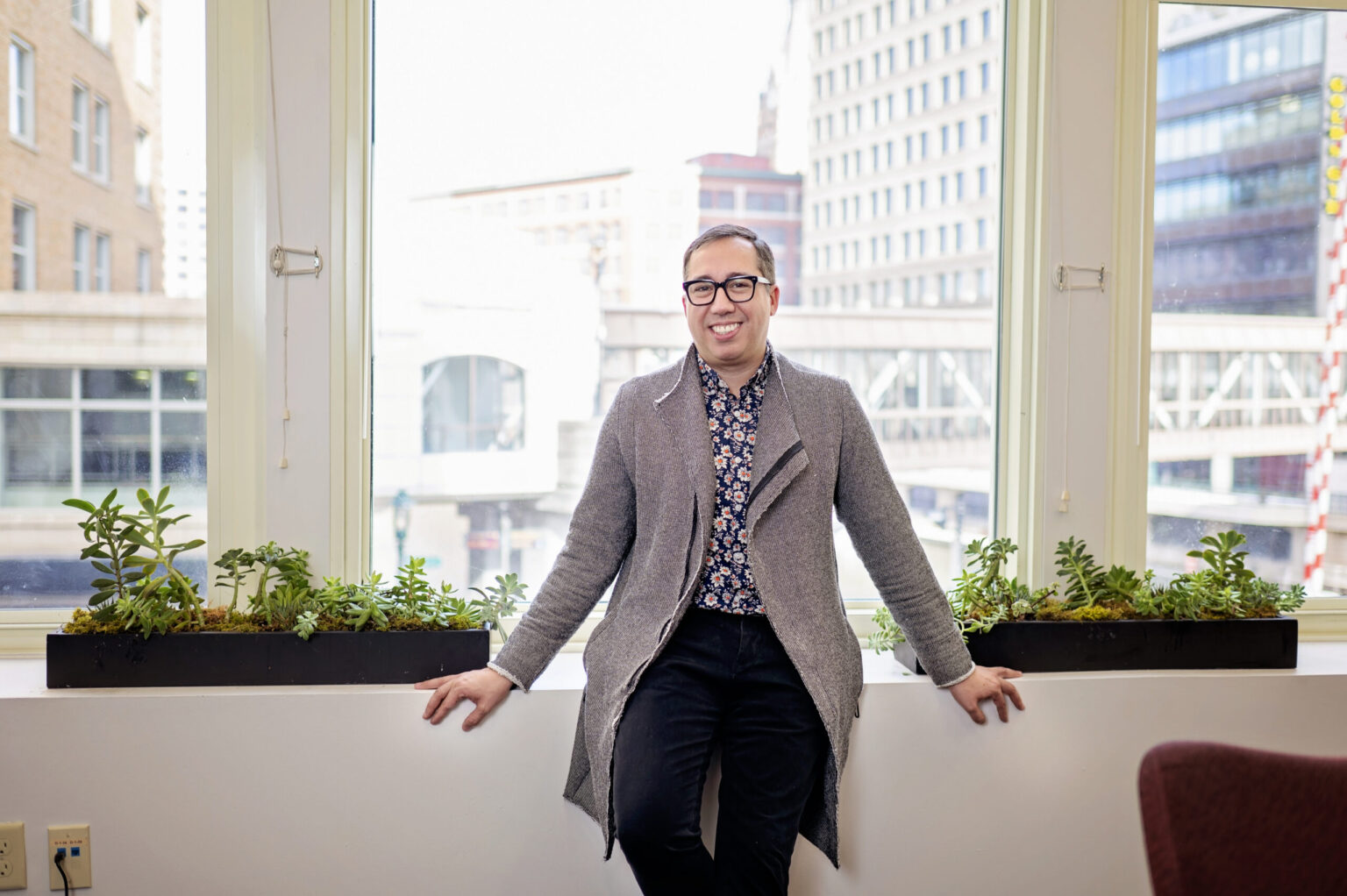 A person wearing glasses and a gray coat sits on a windowsill between two planters, with tall city buildings visible through large windows behind them.