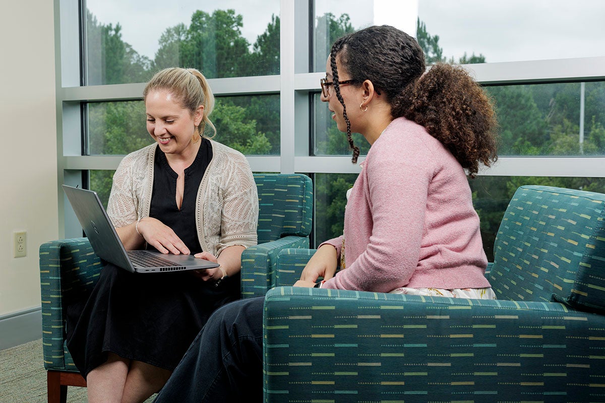 A smiling woman with a laptop sits in a large, plush chair angled close to another woman in a similar chair who is looking at the laptop.