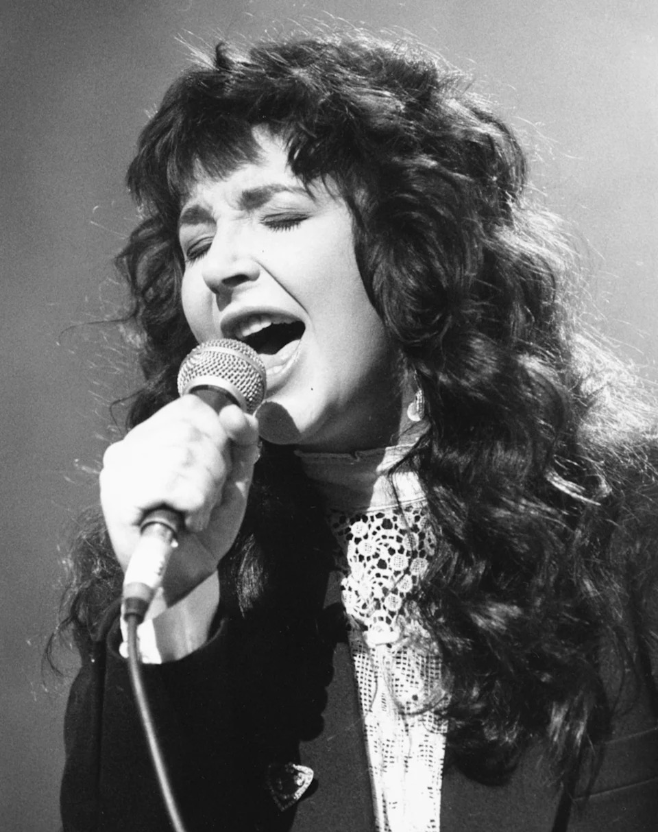 Person singing passionately into a microphone, wearing a lace top under a blazer. They have long, curly hair and eyes closed while performing