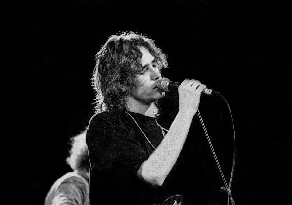 Singer performs passionately on stage, gripping microphone with closed eyes conveying emotion. Casual style, long hair, focused on music