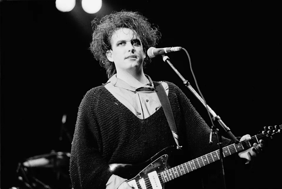 Musician with styled hair plays guitar and sings into a microphone on stage, wearing a loose-fitting sweater over a collared shirt
