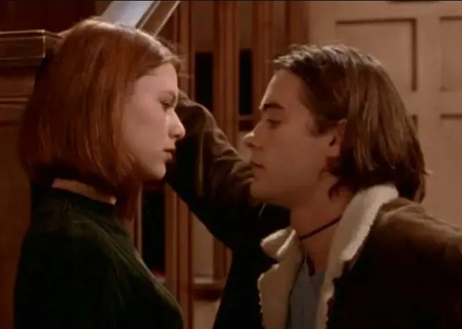 Two young adults stand close, facing each other. The woman looks thoughtful, while the man leans against the wall, appearing intent
