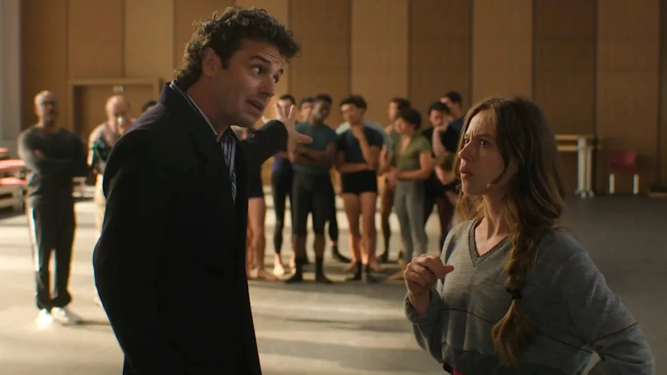 Man and woman converse in a rehearsal space, surrounded by people in dance attire, suggesting a theatrical or performance setting