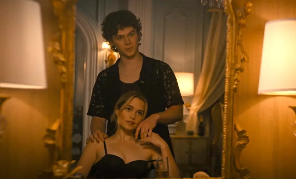 Two people are reflected in a mirror; one is seated, wearing a strapless top, while the other stands behind, with curly hair and a lace shirt