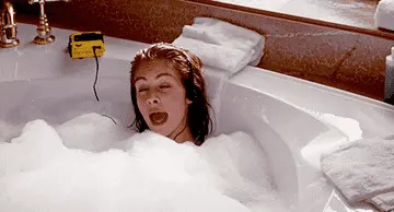 Person in bathtub playfully sticking out tongue amid bubbles, with a yellow radio on the tub's edge
