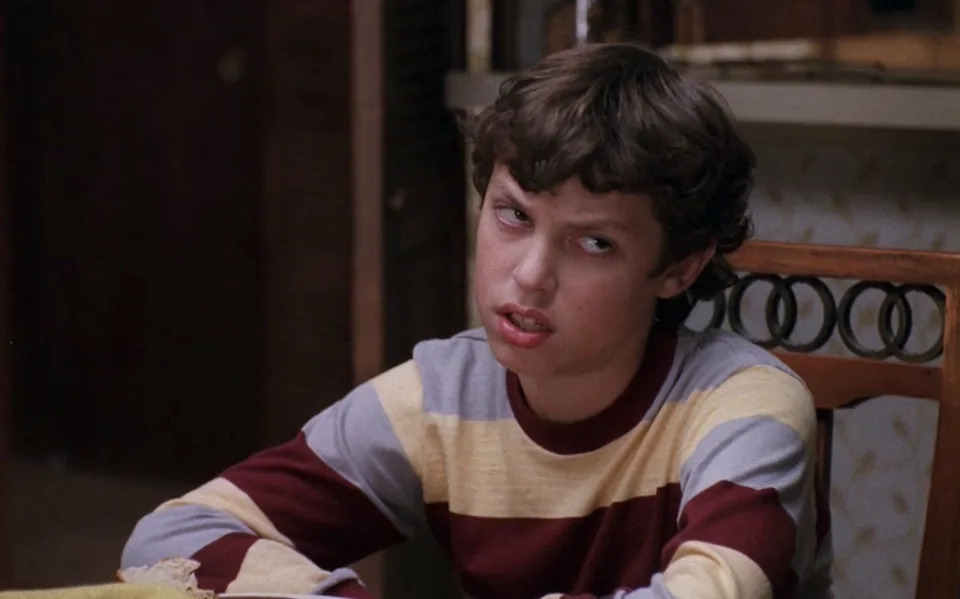 A person with curly hair looks up with a skeptical expression, wearing a striped sweater, seated at a table indoors