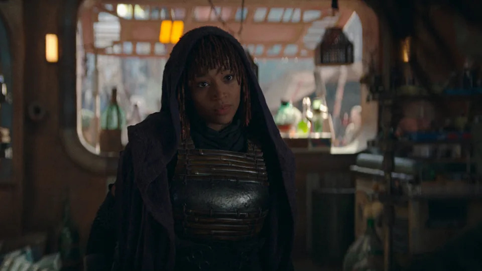 Person in futuristic attire with a dark, textured cloak in a sci-fi setting with shelves and objects in the background