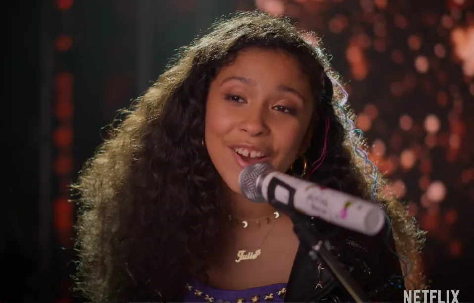 A person sings into a microphone on stage, wearing a necklace and a studded jacket