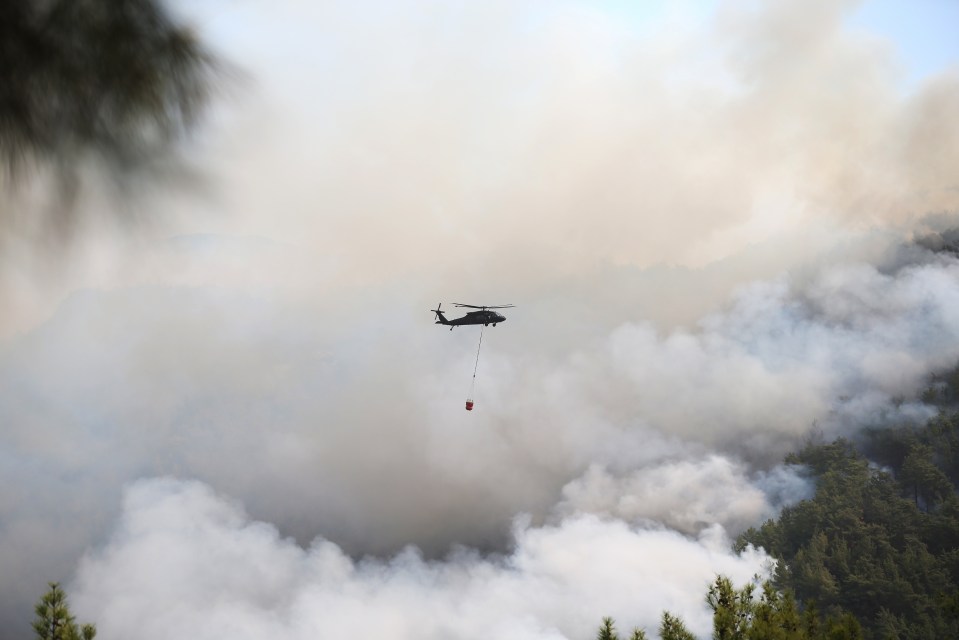 A helicopter with a water bucket flies above a smoky forest wildfire.