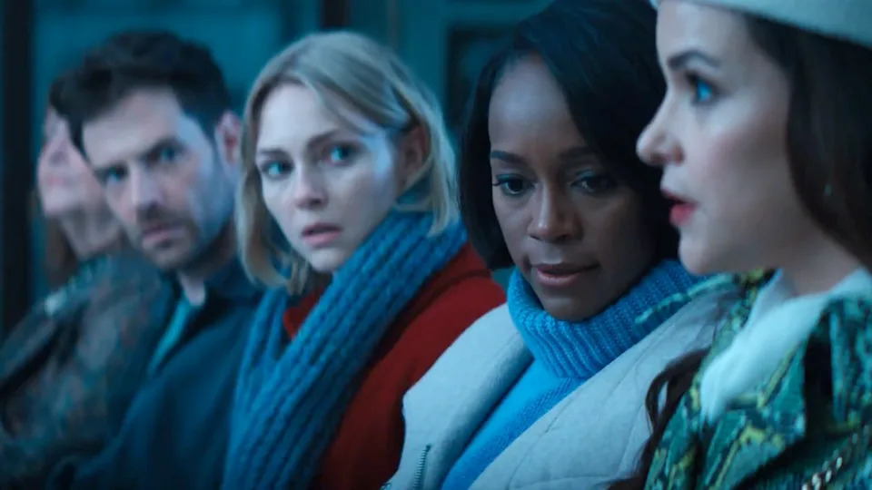 A group of solemn people, seated, looking towards a woman in a green-patterned coat