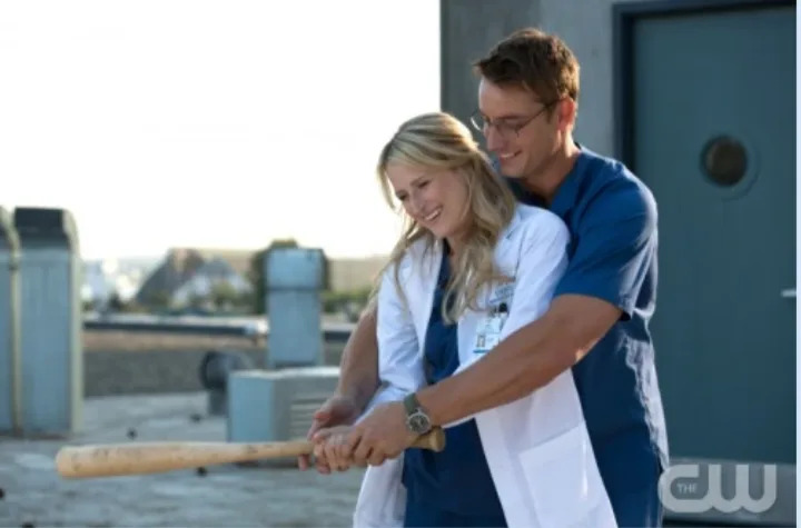 Two people in medical scrubs and a white coat share a moment on a rooftop. One helps the other hold a baseball bat, both smiling