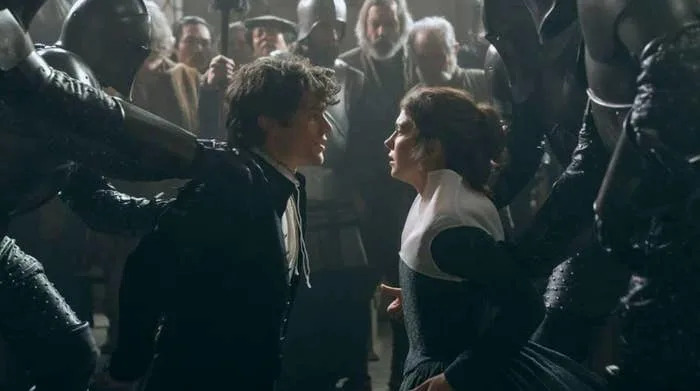 Two actors in period costumes face each other intensely amidst armored figures, while onlookers watch. Scene suggests dramatic tension