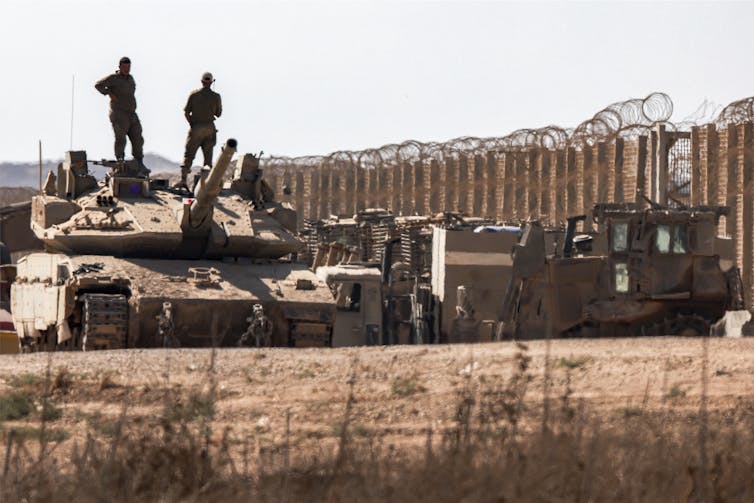 Two men stand on top of a tank with a large fence behind them.