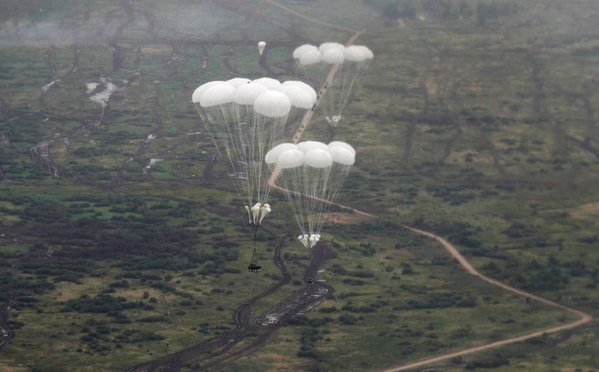 In a photo taken from a nearby aircraft, several parachutes that are white are shown above a lightly populated land mass.