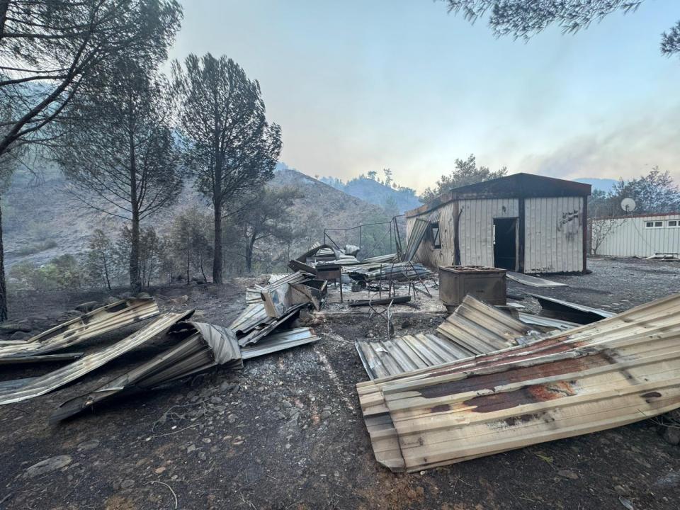 Aftermath of a forest fire showing a burnt house and damaged barns, with charred trees and a smoky sky in the background.