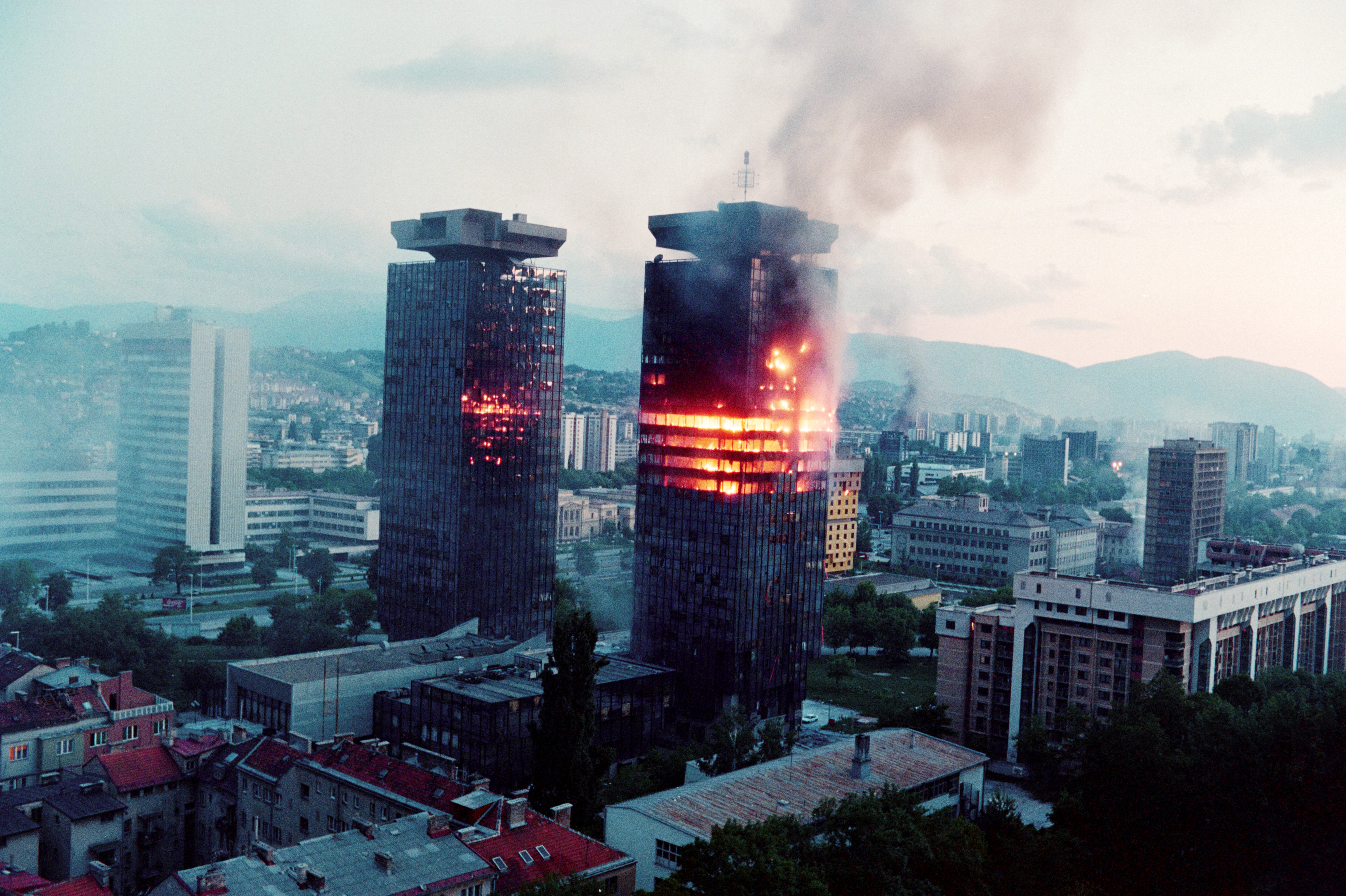 Two twin towers in Sarajevo burning, with fire and smoke billowing from multiple floors, surrounded by other city buildings and distant mountains.