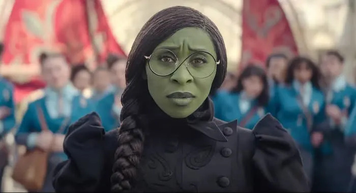 Cynthia Erivo dressed as Elphaba. Her skin is green and she is wearing glasses and a black dress while her hair is in a braid